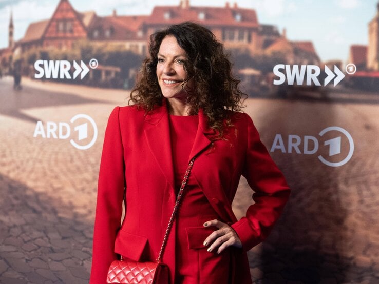 Christine Neubauer mit dunklen lockigen Haaren trägt ein elegantes rotes Outfit, dazu goldenen Nagellack und eine passende rote Tasche vor einem städtischen Hintergrund.
