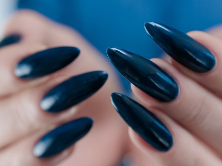 Nahaufnahme von Händen mit langen, glänzend dunkelblau lackierten, spitz zulaufenden Fingernägeln vor unscharfem, blauem Hintergrund.