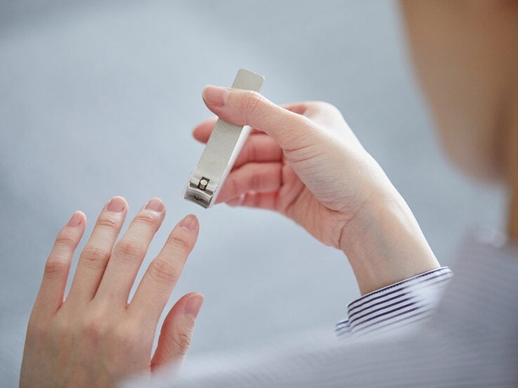 Eine Person mit gestreifter Manschette hält einen silbernen Nagelknipser und schneidet Fingernägel vor neutral grauem Hintergrund.