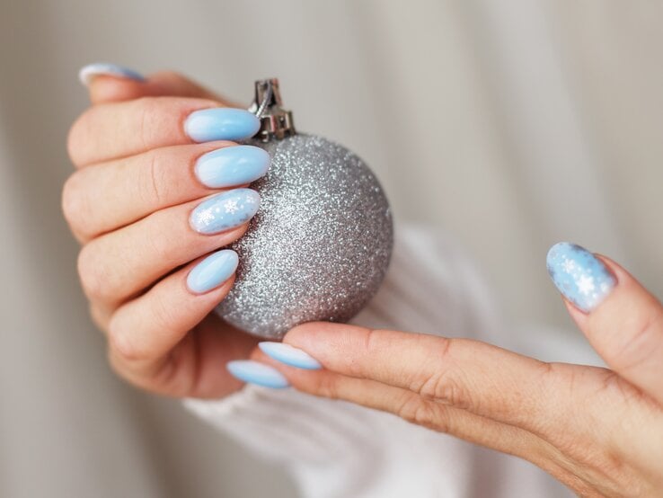 Hände mit hellblau lackierten, mandelförmigen Nägeln, ein Nagel mit weißen Schneeflocken, hält eine silberne Weihnachtskugel.