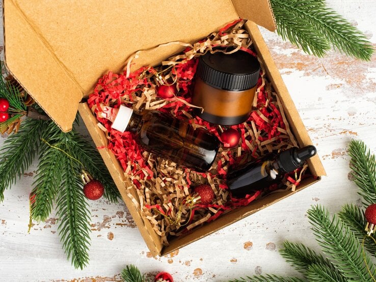 Kosmetik-Set in brauner Box mit rotem Füllmaterial, dekoriert mit Tannengrün und festlichen Christbaumkugeln.