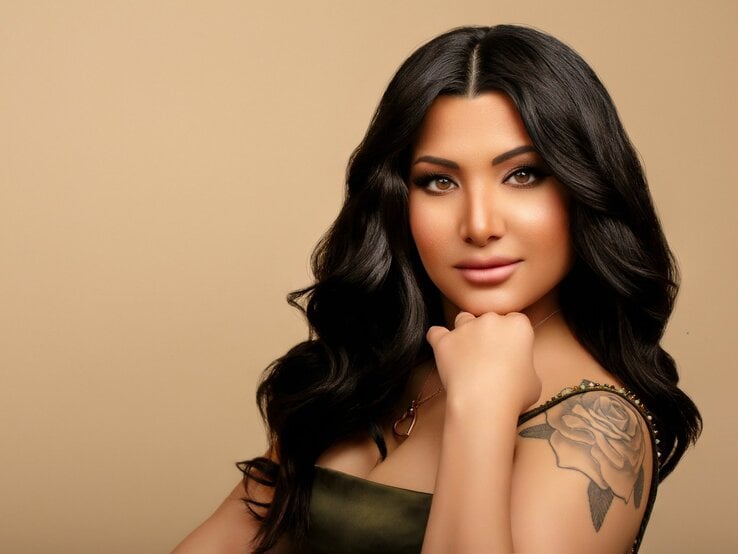 Frisörin Nesrin Obermeier mit langen, gewellten, dunklen Haaren und Make-up. Sie hat ein herzförmiges Gesicht, volle Lippen und große Augen. Auf ihrem Oberarm ist ein Tattoo einer Rose. Sie trägt ein olivgrünes Kleid. Der Hintergrund ist in einem warmen Beigeton gehalten. | © Nesrin Obermeier