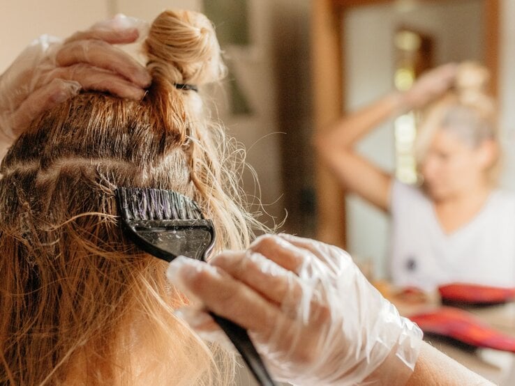 Frau färbt sich ihre Haare vor dem Spiegel mithilfe eines Pinsels, sie trägt Einmalhandschuhe