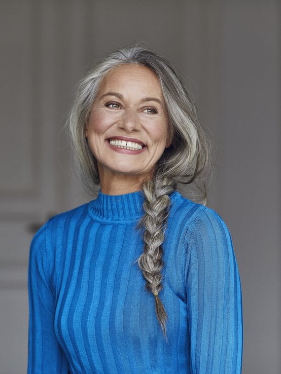 Eine lächelnde ältere Frau mit langen grauen Haaren im Zopf trägt ein blaues Kleid und steht vor einem neutralen, lichtdurchfluteten Hintergrund. | © Getty Images/ Westend61