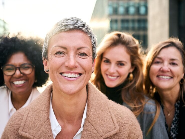 Eine Frau mit kurzem grauem Haar und beigem Mantel lächelt in die Kamera, drei Freundinnen lachen im Hintergrund.
