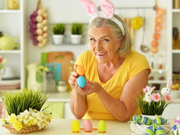 Eine ältere Frau mit grauen Haaren und Hasenohren-Stirnband hält bunt gefärbte Ostereier und lächelt fröhlich in einer frühlingshaft dekorierten Küche.