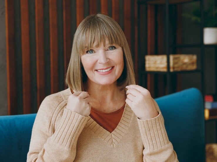 Lachende ältere Frau mit kinnlangem, blondem Bob und Pony sitzt im beigen Pullover auf blauem Sofa vor Holzpaneelen.