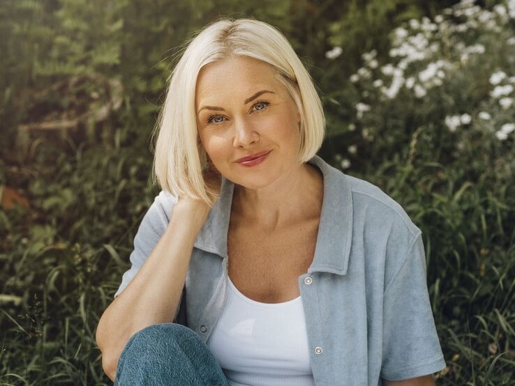 Frau mit kurzem, blondem French Bob und entspannter Miene sitzt im Gras, umgeben von weißen Blüten und Gräsern.
