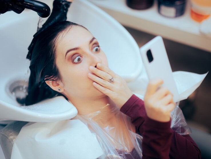 Junge Frau mit dunklem Haar hält schockiert das Handy hoch, während ihr beim Friseur die Haare gewaschen werden.