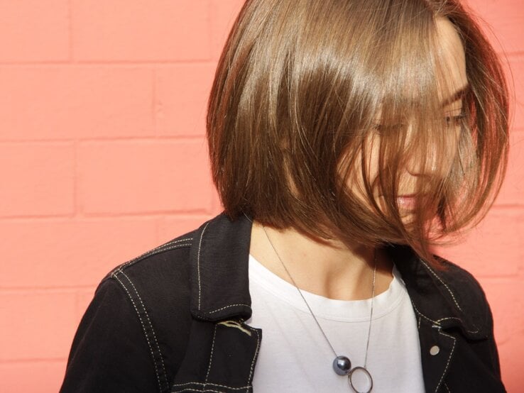 Eine junge Frau mit schulterlangem, braunem Bob trägt ein weißes T-Shirt und eine schwarze Jacke, ihr Blick ist nach unten gerichtet vor pinkem Hintergrund.