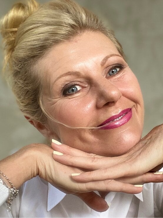 Blondhaarige ältere Frau (Renate Zott) mit Dutt lächelt sanft, stützt das Kinn auf die Hände und trägt rosa Lippenstift sowie weiße Bluse. | © Renate Zott