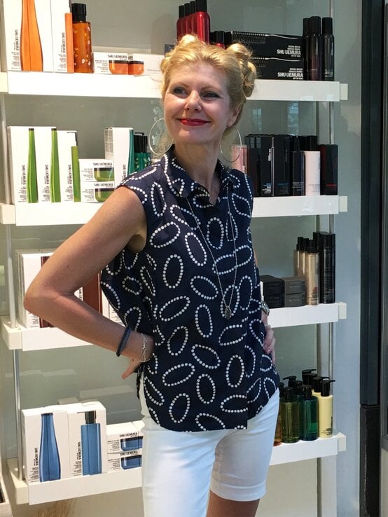 Blondhaarige Renate Zott mit Doppeldutt und rotem Lippenstift posiert fröhlich vor Regal mit Pflegeprodukten in eleganter Sommerkleidung. | © Renate Zott