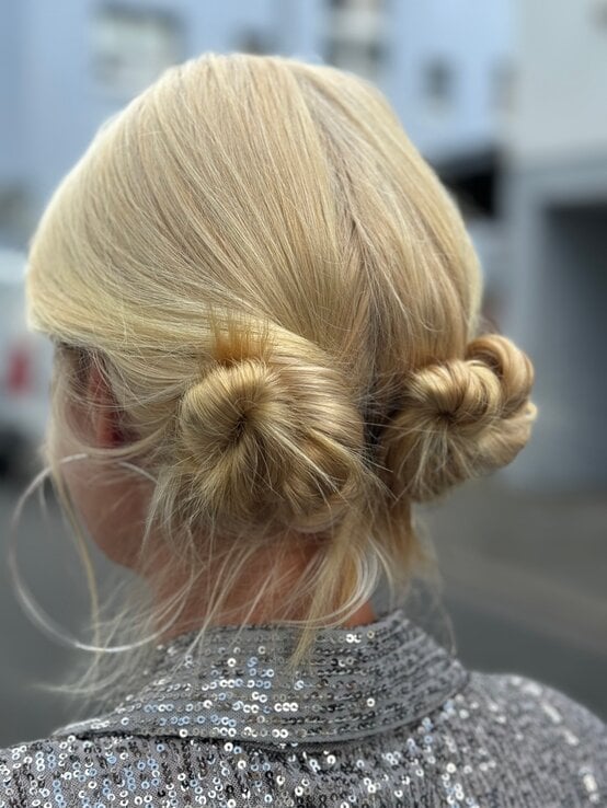 Rückansicht einer blondhaarigen Renate Zott mit zwei tiefen Dutts und glitzerndem Paillettenoberteil vor unscharfem Stadthintergrund. | © Renate Zott