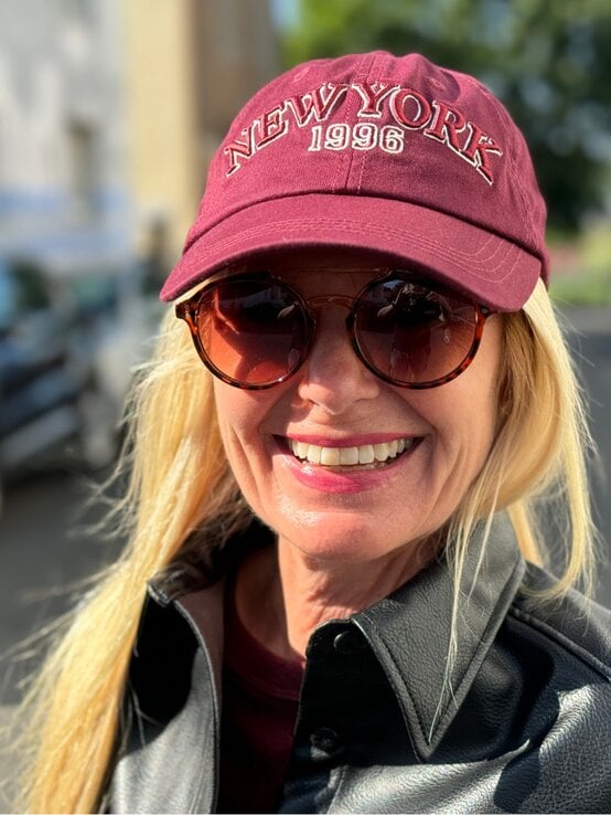 Blondhaarige Renate Zott mit bordeauxfarbener New-York-Kappe und Sonnenbrille lacht strahlend in der Sonne auf einer Stadtstraße. | © Renate Zott