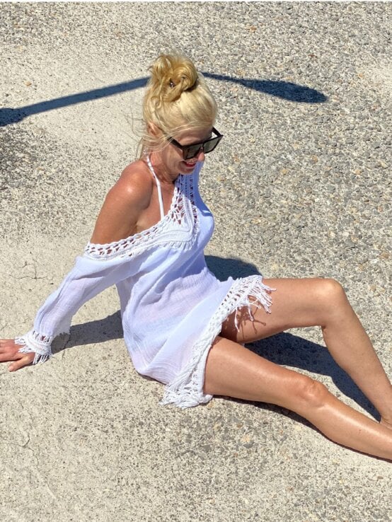 Renate Zott mit Dutt, Sonnenbrille und gehäkelter Tunika genießt barfuß die Sonne auf heller Steinfläche neben Felsen und Wasser. | © Renate Zott