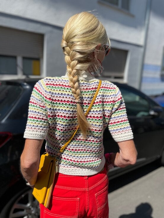 Blondhaarige Renate Zott mit geflochtenem Zopf trägt buntes Häkelshirt, rote Hose und gelbe Tasche im Sonnenlicht auf der Straße. | © Renate Zott