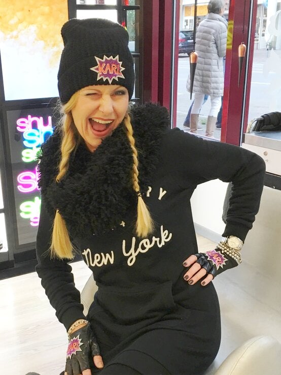 Lachende Renate Zott mit blonden Zöpfen, Statement-Mütze und Handschuhen zeigt sich augenzwinkernd in trendigem Winteroutfit im Geschäft. | © Renate Zott