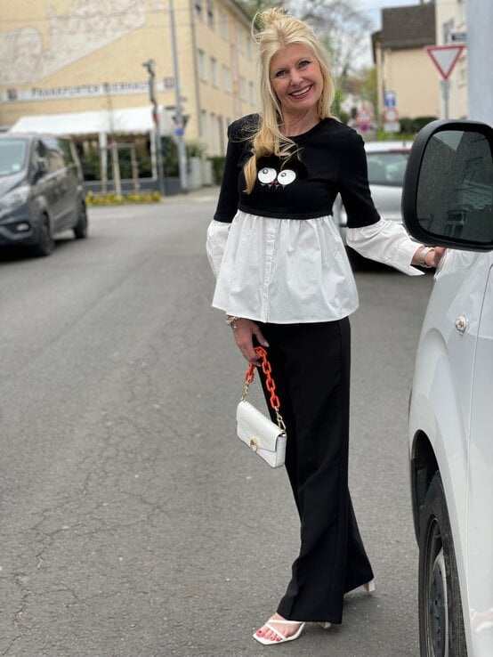 Modebewusste Renate Zott mit weißer Handtasche, schwarzer Hose und weißen Sandaletten posiert fröhlich in urbaner Umgebung. | © Renate Zott