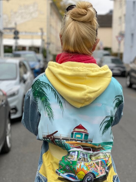 Rückansicht von Renate Zott mit klassischem Dutt, farbenfrohem Kapuzenpulli und Jeansjacke im urbanen Umfeld bei bedecktem Himmel. | © Renate Zott