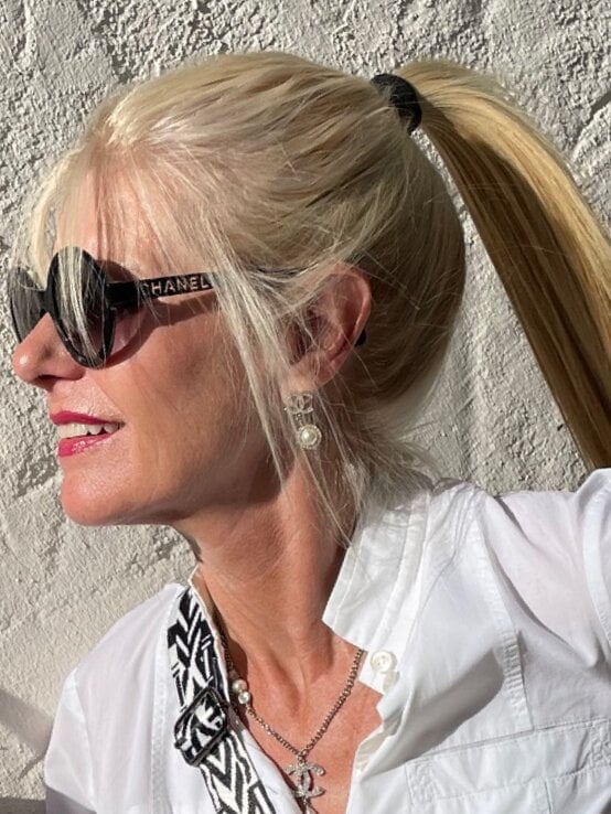 Blondhaarige Renate Zott mit hohem Pferdeschwanz und Chanel-Sonnenbrille blickt lächelnd in die Sonne vor rauer weißer Wand. | © Renate Zott