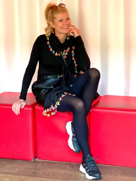 Modebewusste Renate Zott mit Pferdeschwanz, Strumpfhose und farbenfrohem Detail-Look sitzt locker lächelnd auf roten Würfeln im Innenraum. | © Renate Zott