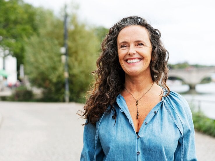 Zu sehen ist eine lächelnde Frau mittleren Alters, die lockiges Haar hat.