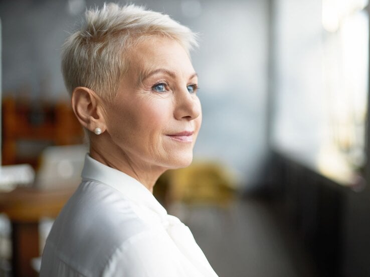 Reife Frau mit stylischer Pixie-Frisur - einer Kurzhaarfrisur für dünnes Haar - und Perlenohrringen trägt weiße Bluse und schaut verträumt ins Tageslicht.