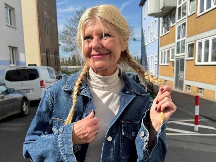 Renate Zott mit geflochtenen Zöpfen, blauer Jeansjacke und hellen Lippen lächelt sanft bei Sonnenschein zwischen Wohnhäusern.