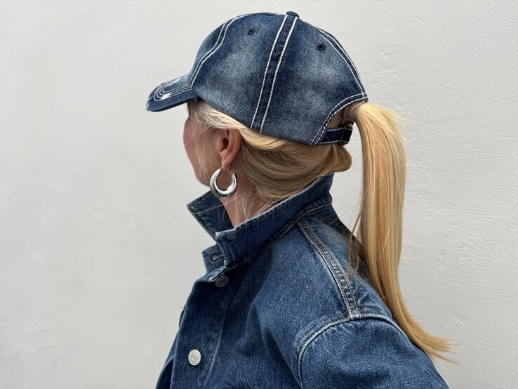 Blondhaarige Renate Zott mit Pferdeschwanz und silbernen Creolen trägt Jeansjacke und Baseballcap, im Profil vor grauer Wand.