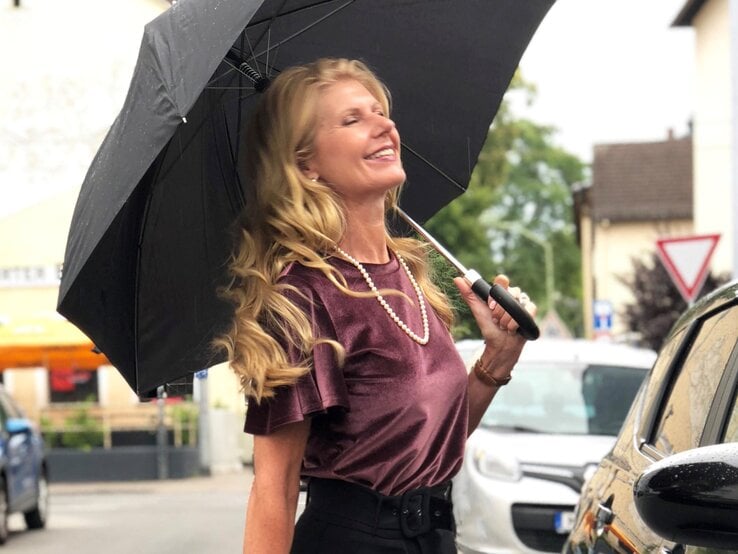 Renate Zott mit Perlenkette und violetter Bluse hält fröhlich einen schwarzen Schirm in regennasser Straße.