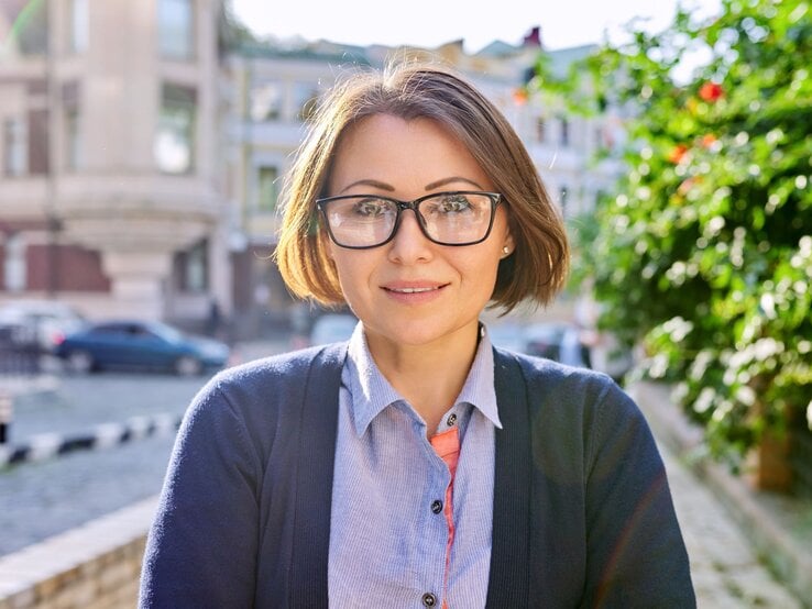 Freundlich blickende Frau mit braunem Bob und schwarzer Brille steht bei Sonnenschein in einer ruhigen Stadtstraße.