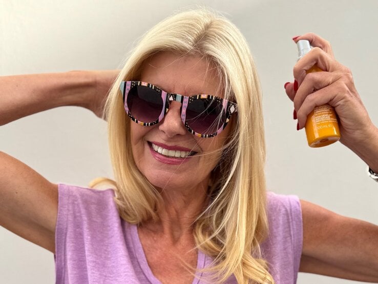 Renate Zott mit offenem blondem Haar posiert in pinker Sonnenbrille und lila Shirt, hält Sprayflasche hoch.