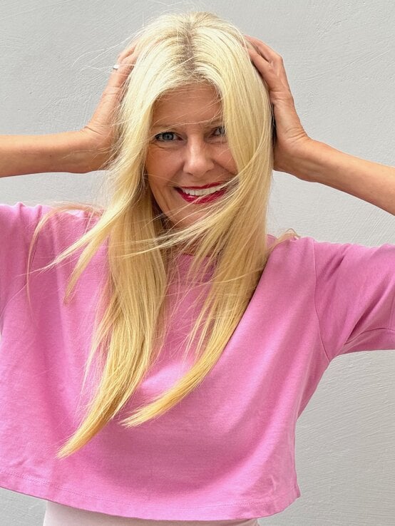Renate Zott mit langen blonden Haaren und pinkem T-Shirt lächelt lebhaft, hält sich die Haare vor hellgrauer Wand. | © Renate Zott