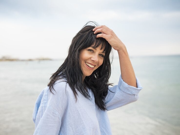 Lachende dunkelhaarige Frau mittleren Alters in hellblauer Bluse streicht sich am Strand fröhlich das Haar aus dem Gesicht.