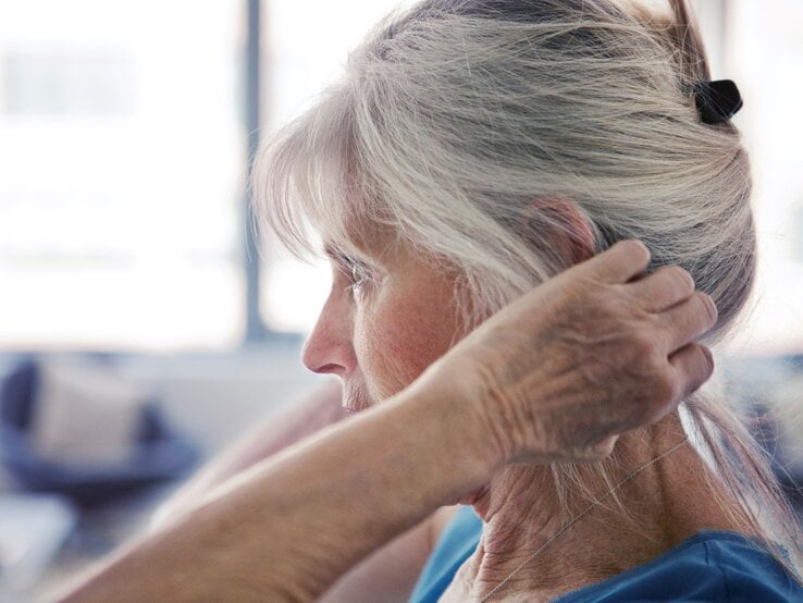 Ältere Frau mit grauen Haaren bindet sich konzentriert die Haare zusammen, seitlich im Profil vor heller Fensterkulisse.