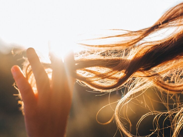 Nahaufnahme einer Hand, die durch langes Haar gleitet, das in der warmen Abendsonne leuchtet und sich im Wind bewegt.