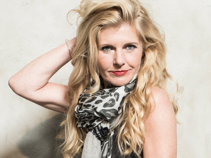 Frau mit langen, welligen blonden Haaren trägt einen schwarz-weiß gemusterten Schal vor beigem Hintergrund und hebt eine Hand ins Haar. | © Renate Zott 