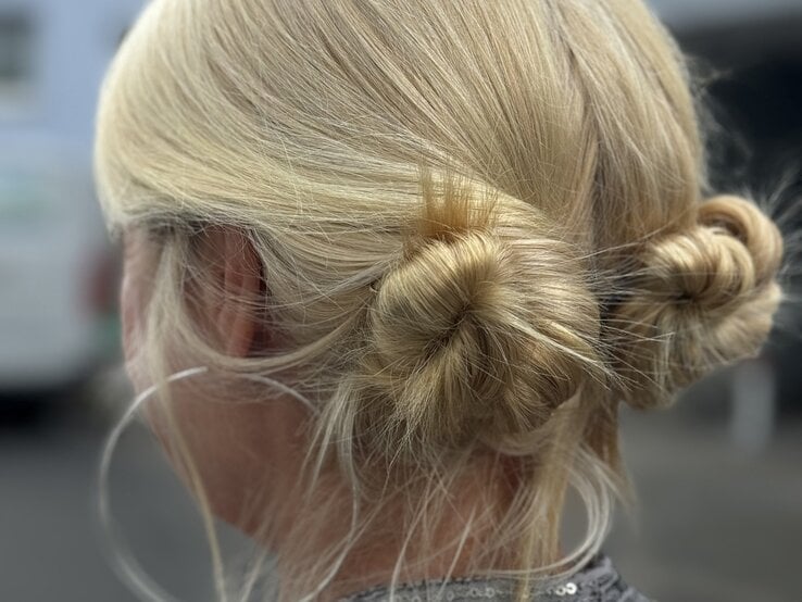 Zwei blonde Haarknoten, kunstvoll nebeneinander gesteckt, ergänzt durch schlichte Kreolen vor einem dezent urbanen Hintergrund. | © Renate Zott 