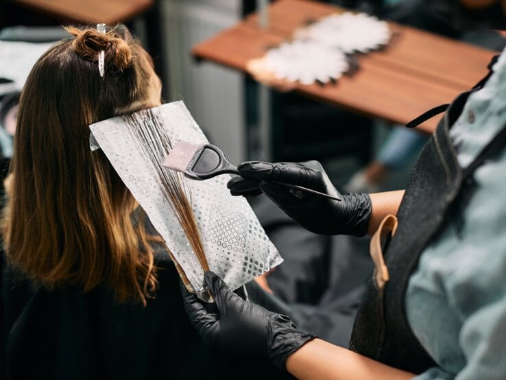 Friseur mit schwarzem Handschuh trägt mit Pinsel Haarfarbe auf braune Strähnen einer Frau auf, die in einem Salon sitzt.
