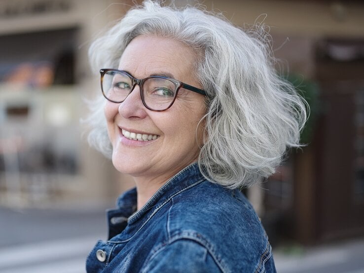 Nahaufnahme von grauem Haar und einer blauen Jeansjacke vor Bokeh-Stadtatmosphäre mit verschwommenen Gebäuden.