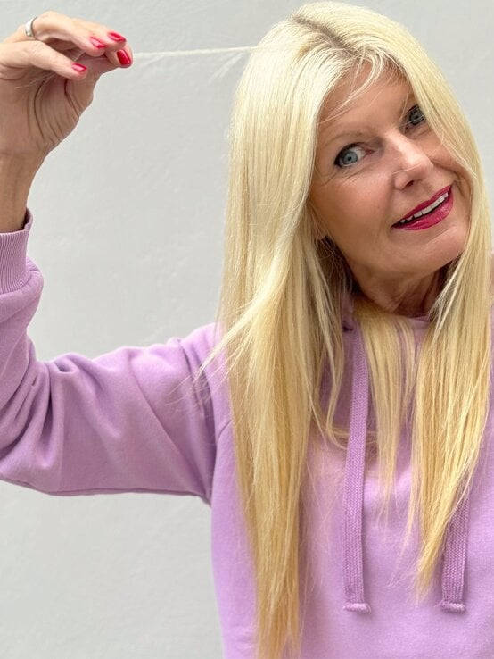 Blondhaarige ältere Frau in fliederfarbenem Hoodie lächelt charmant und hält eine feine Haarsträhne vor hellem Hintergrund. | © Renate Zott