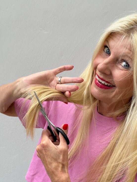 Blondhaarige ältere Frau mit pinkem Lippenstift schneidet lachend eine Haarsträhne mit einer Schere vor grauer Wand. | © Renate Zott
