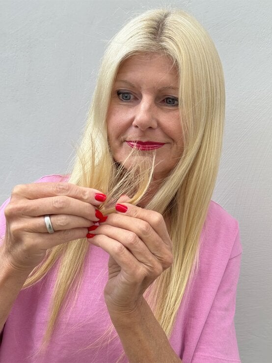 Ältere Frau mit glattem, blondem Haar und pinkem Shirt prüft nachdenklich ihre Haarspitzen vor grauer Wand. | © Renate Zott