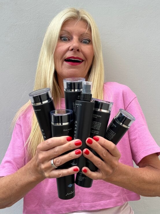 Blondhaarige Frau in rosa Shirt blickt erstaunt auf mehrere schwarze Haarpflegeprodukte, die sie mit beiden Händen hält. | © Renate Zott