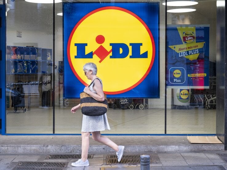 Grauhaarige Frau in weißem Sommerkleid und Sneakers läuft an einer Lidl-Filiale mit großem gelbem Logo vorbei.