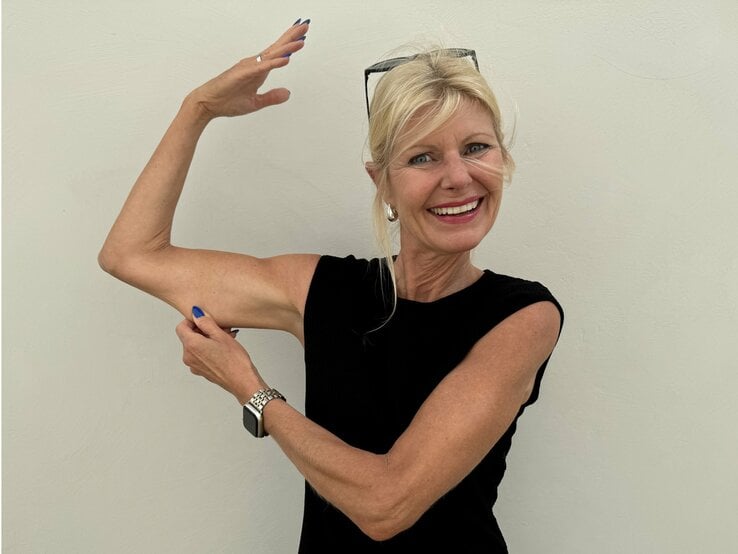 Frau in einem ärmellosen, schwarzen Oberteil. Sie hebt einen Arm und zeigt mit der anderen Hand auf ihren Oberarm, was ihre trainierte und fitte Erscheinung betont. Sie trägt eine Uhr und hat blaue Fingernägel.