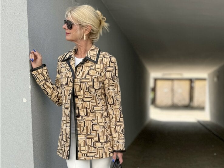 Frau mit blonden Haaren, die zu einem lockeren Knoten hochgesteckt sind. Sie trägt eine Sonnenbrille und eine gemusterte Jacke in Beige- und Brauntönen mit geometrischen Mustern. 