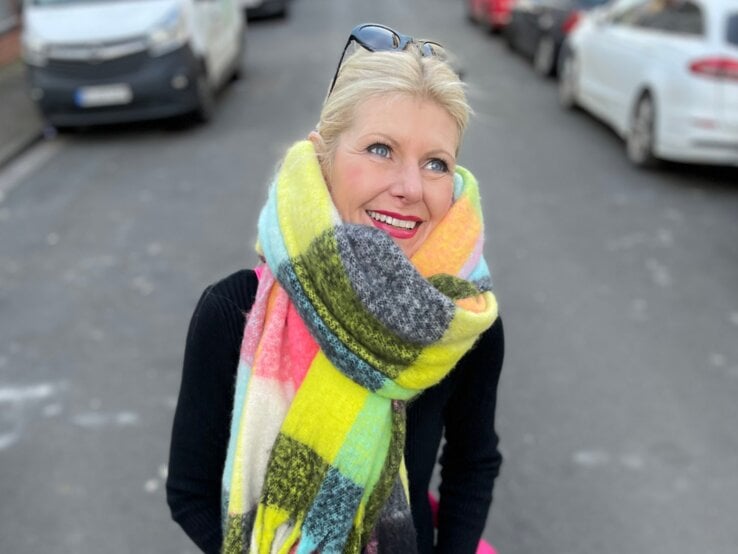 Renate Zott mit hochgestecktem Haar, buntem XXL-Schal und schwarzem Outfit steht auf einer Stadtstraße mit geparkten Autos.
