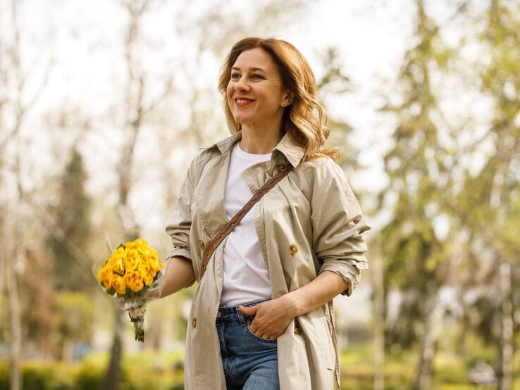 Zu sehen ist eine lächelnde Frau, die Rosen in der Hand hat und einen Trenchcoat trägt.