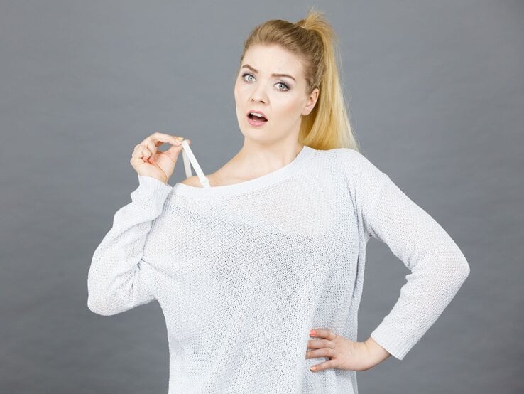 Junge blonde Frau mit Pferdeschwanz zieht überrascht den Träger ihres weißen Pullovers herunter, grauer Hintergrund.